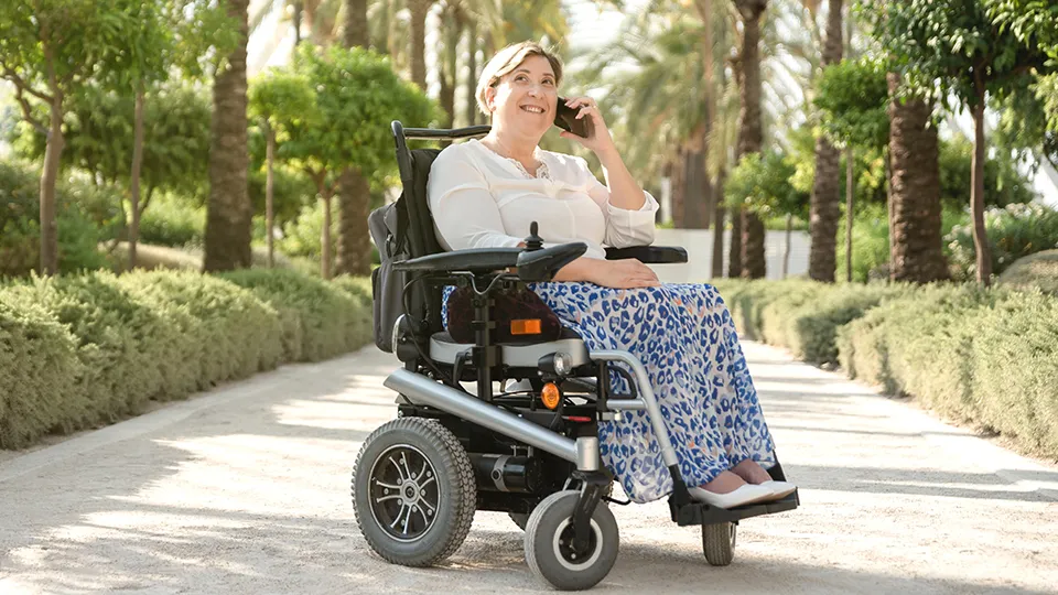 A smiling woman in a power wheelchair enjoys a sunny day in a tree-lined park, talking on her phone. She wears a long blue patterned skirt and a white blouse, surrounded by lush greenery and palm trees.
