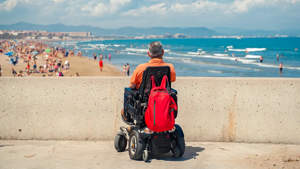 A man in a power wheelchair faces the ocean while sitting on a beach promenade, with a red backpack hanging from the chair. A crowded beach and rolling waves stretch into the distance under a partly cloudy sky.